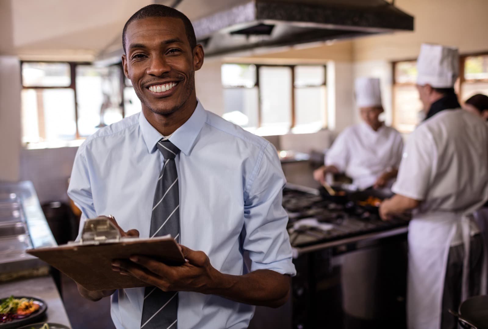 An inspector with a clipboard in a commercial kitchen as chefs work at the line behind him.
