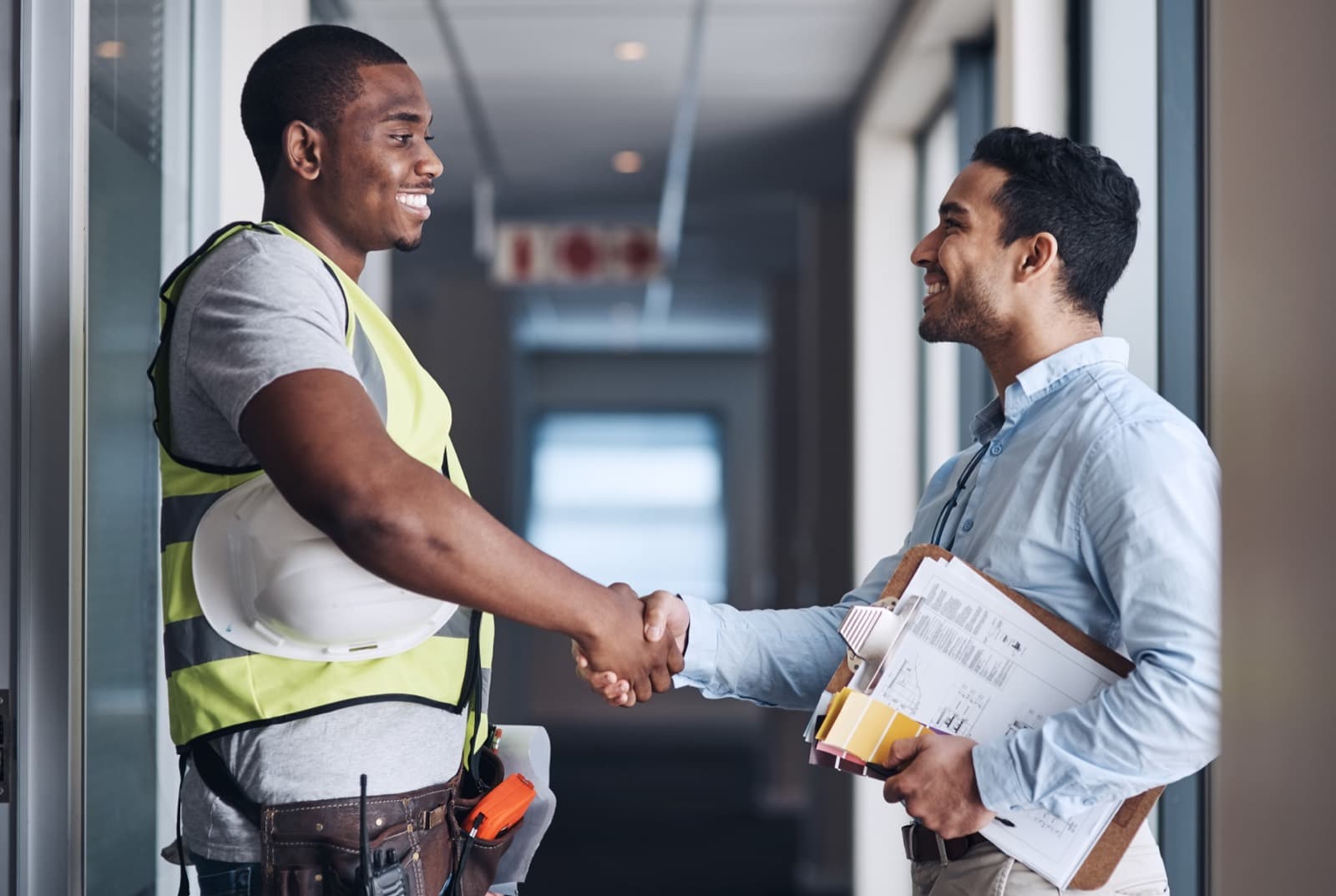 A contractor and a client shaking hands over project plans in a commercial building.